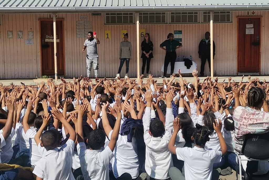The backs of a group of elementary school students sitting cross-legged and facing five adults in a school yard. The students are raising their hands as if answering a question